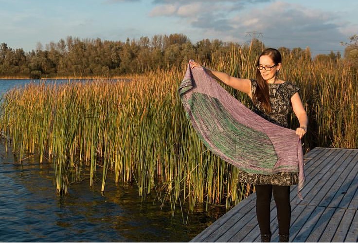 A woman, the designer, is showing off her shawl. It's the Mary Shelley shawl. She is standing in front of a lake with the sun shining behind her.